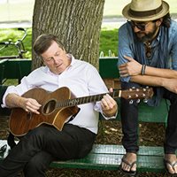 Photo of Mayor John Tory sitting on a park bench playing a guitar. A man sits next to him and watches. 