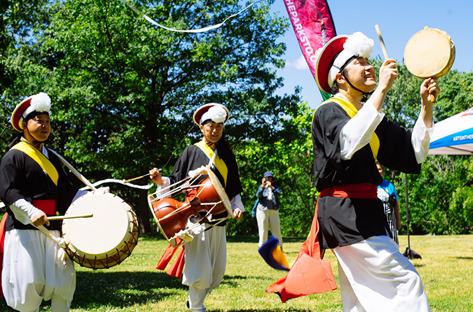 A photo of three musicians from Ensemble Jeng Yi. They are wearing traditional Korean drumming costumes and carrying drums that they are playing.