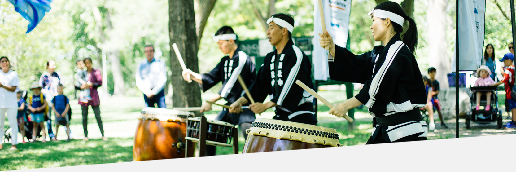 A photo of of three Japanese Taiko drummers in a park. Onlookers watch. 