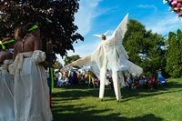 A performer wearing all white stretches their arms while on stilts