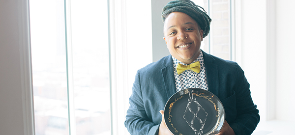 A photo of Syrus Marcus Ware standing beside a window, smiling, wearing a dark jacket, bowtie and patterned shirt. They hold a black plate in both hands, with a gold and white design 