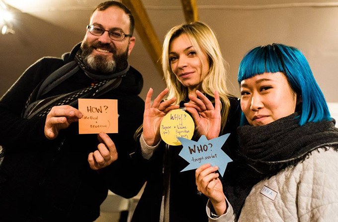 A trio holds up different paper cut outs. Each cut out has a different word on it: 'How,' 'Why,' 'Who?'