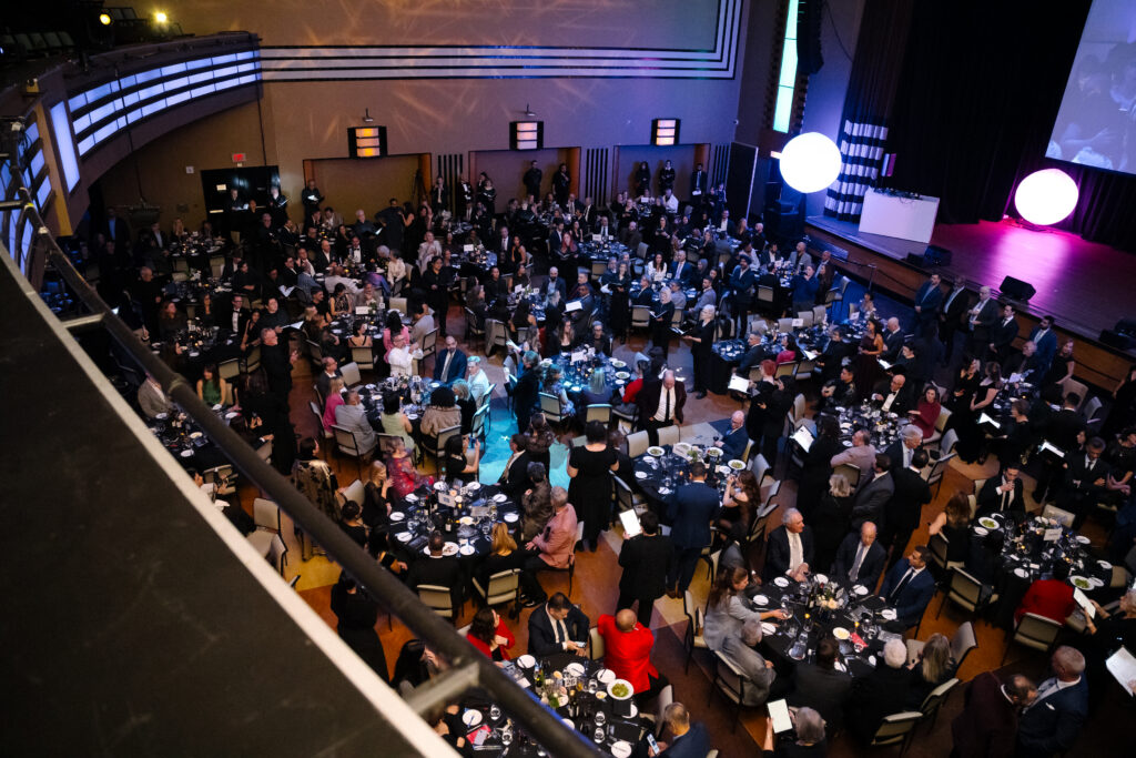 Many people pictured from above. They are seated at elegant dinner tables in low lighting. Colourful orbs light the room.