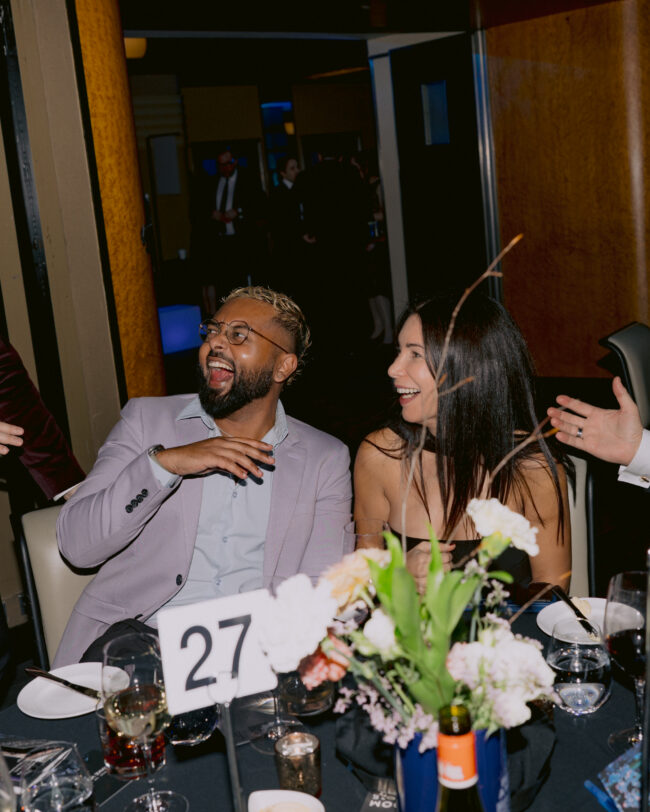 Four people sit at a dinner table in suits and dresses, talking and laughing.