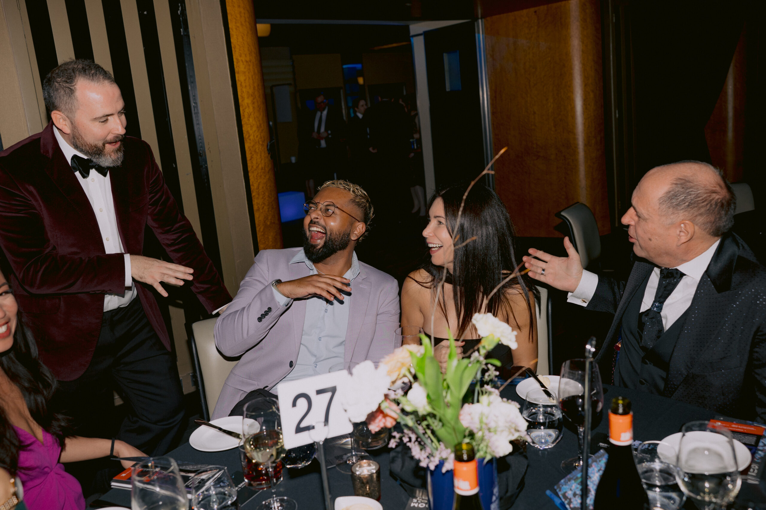 Four people sit at a dinner table in suits and dresses, talking and laughing.