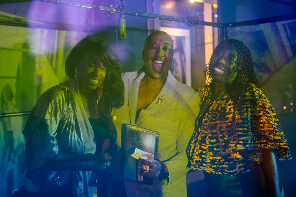 Three friends smile inside a colourful light installation, which makes them appear green, blue, yellow, and purple.