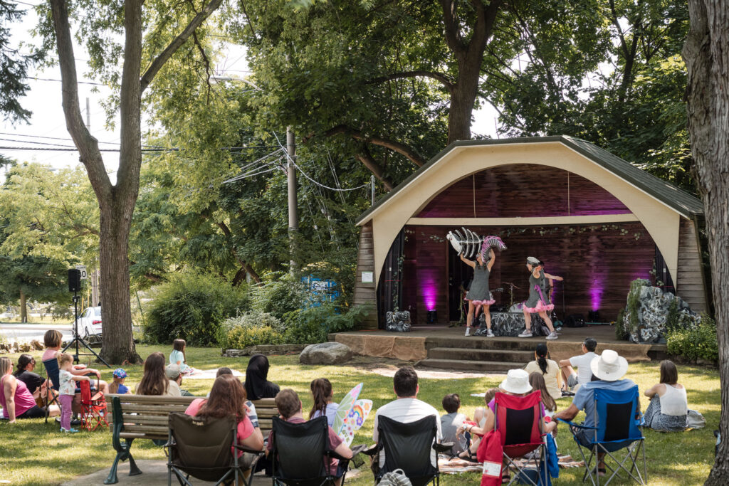 Audience members sit in the park on mats and lawn chairs to view the performance. Two performers onstage hold a large set of fish bones. Photo by Kat Rizza