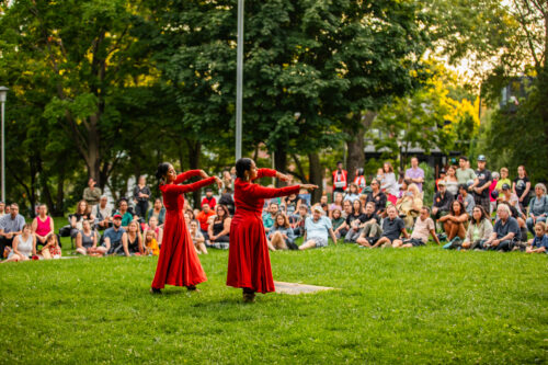 Two dancers dressed in red dance for a crowd in a green park.