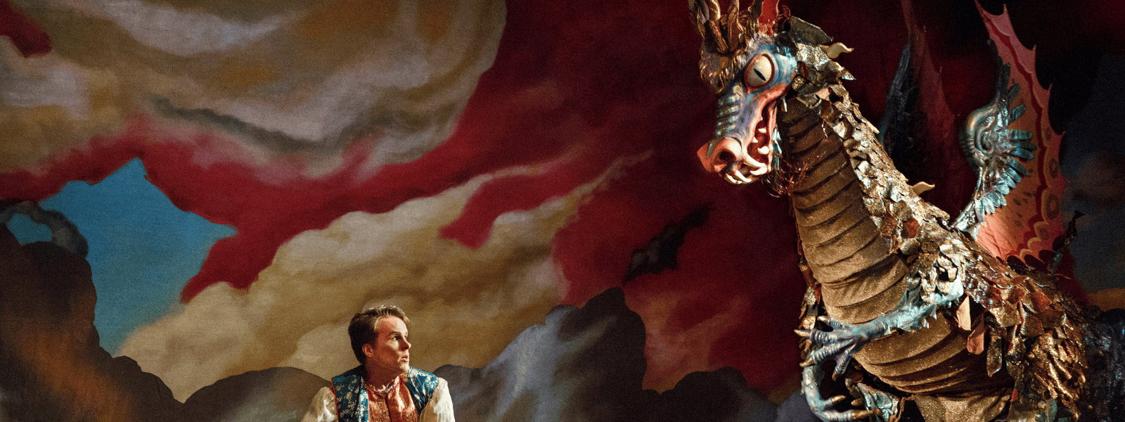 A dramatic stage scene with stormy red and grey clouds painted as a backdrop. On the left, an actor in a colorful vest looks up in awe or concern. On the right, a large, elaborate dragon puppet with shimmering scales, blue and gold wings, and fierce eyes looms over him.