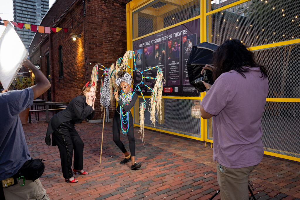 A photographer is doing a photoshoot and posing for him are dancers performers dressed in their costumes with Soul Pepper Theatre ad in the background