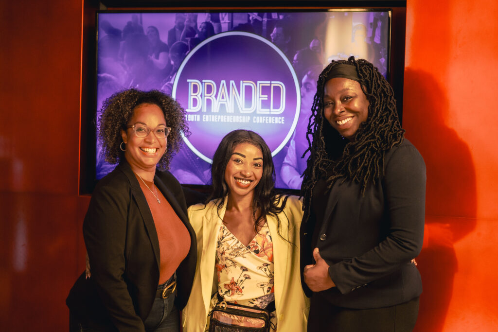 Three women posing for the picture in front of a poster for an event named Branded
