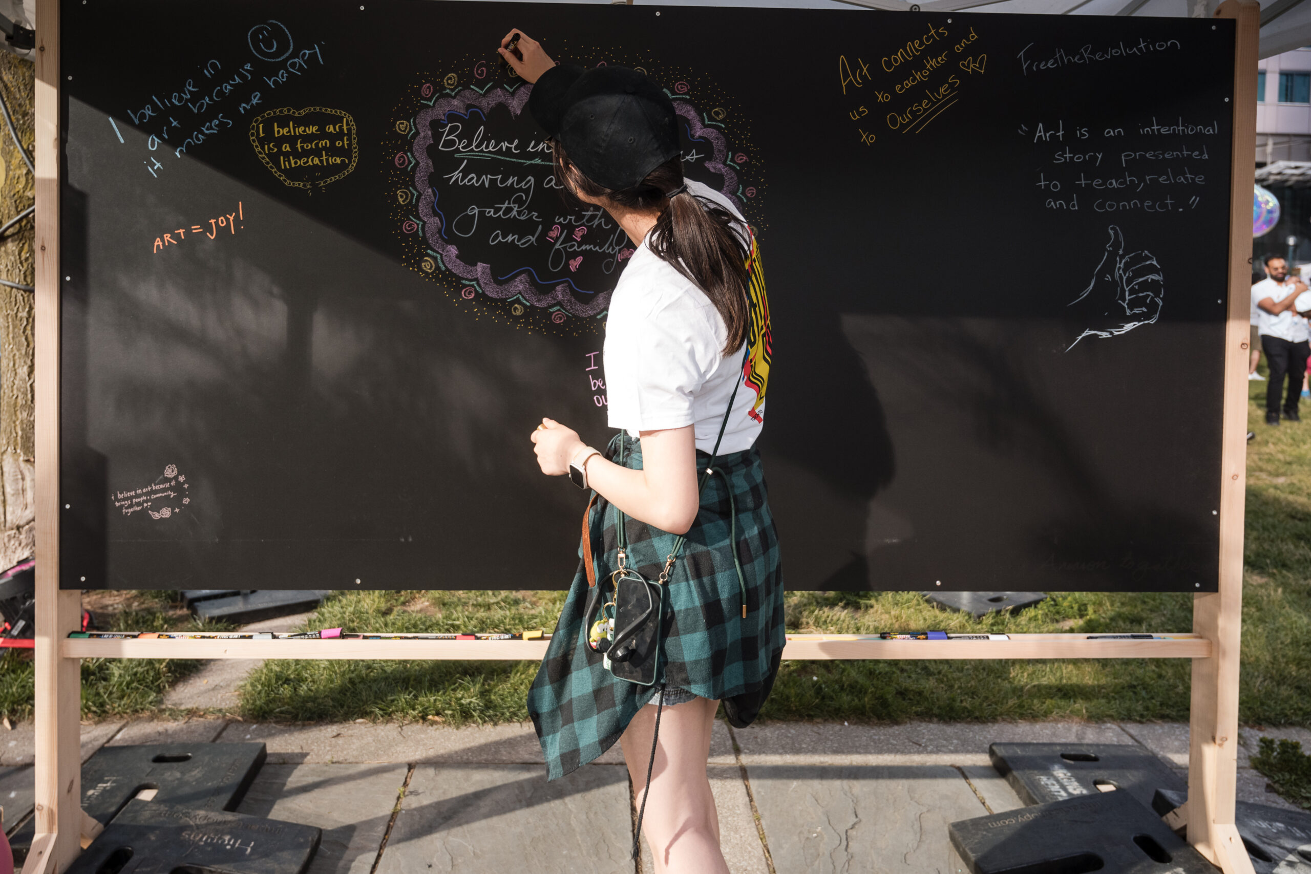 A person writes why they believe in art on a chalkboard filled with other responses.