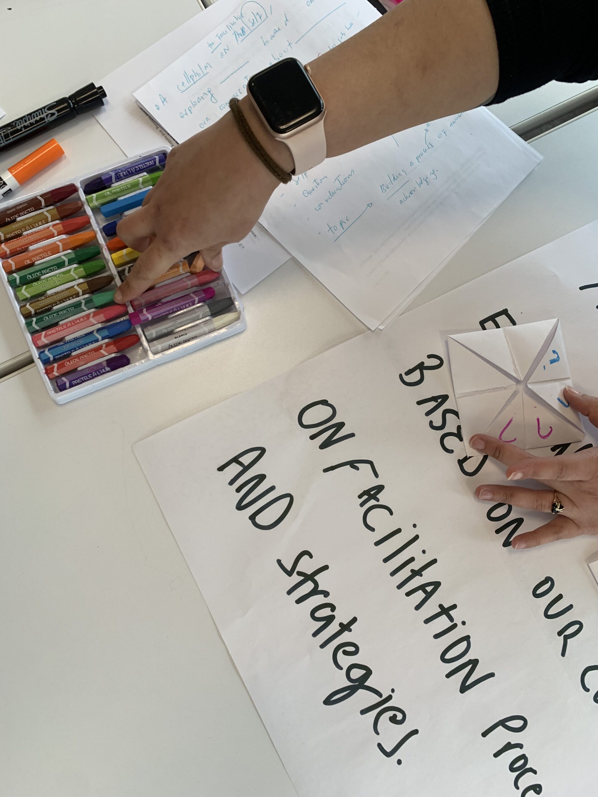 a hand selecting a crayon from a full box of crayons placed on a table with large white paper spread across and one part of the paper seen with writing on it