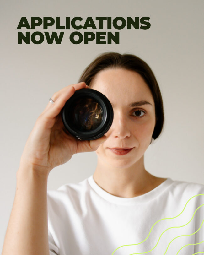 image shows a person holding a camera lens over one of her eye wearing a white t-shirt and looking at you - with the caption "applications new open"