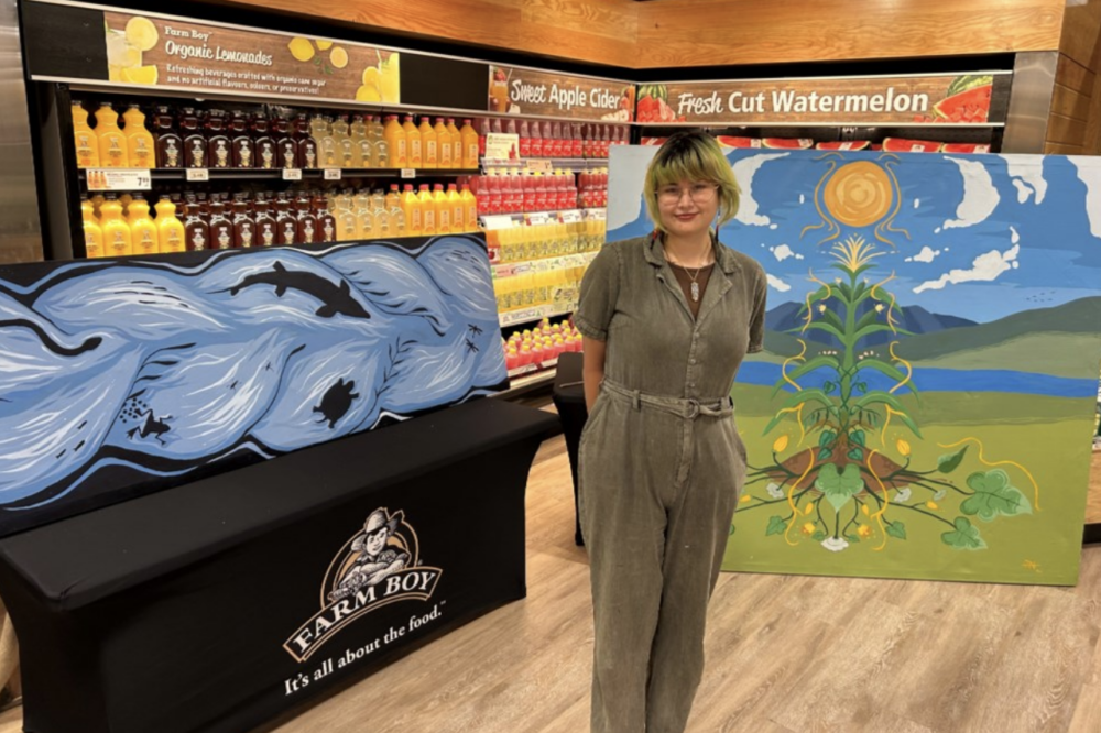 Brenna Nanie stands in a Farm Boy store in front of shelves of goods and an original artwork that depicts a scenic landscape with a sun, sky, mountains, water, grass, and plant.