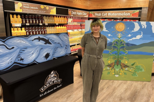 Brenna Nanie stands in a Farm Boy store in front of shelves of goods and an original artwork that depicts a scenic landscape with a sun, sky, mountains, water, grass, and plant.