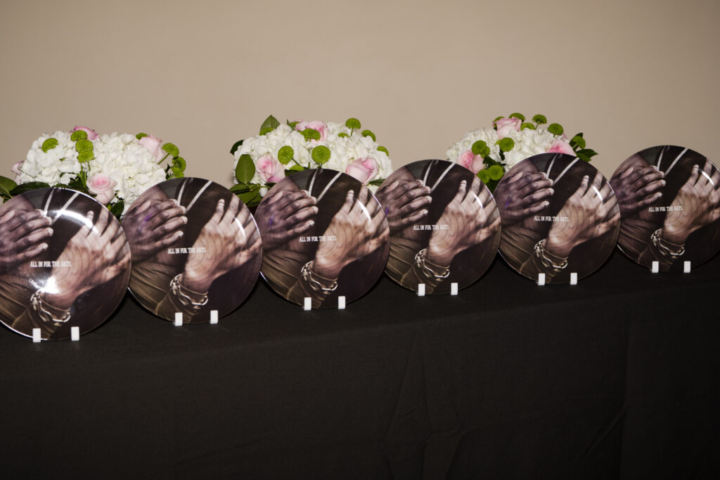 A row of plates stacked vertically on a table. The plate depicts hands touching each other and read "ALL IN FOR THE ARTS".