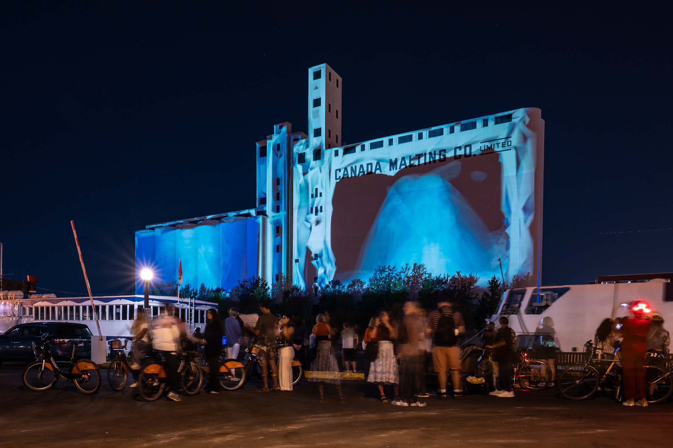 A group of people hang out in front of the silo near the Toronto waterfront at night.
